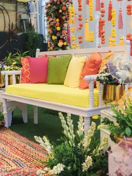 A vibrant and comfortable Mehendi seating area. The setup includes a plush bench with colorful cushions, surrounded by lush greenery and hanging floral tassels.