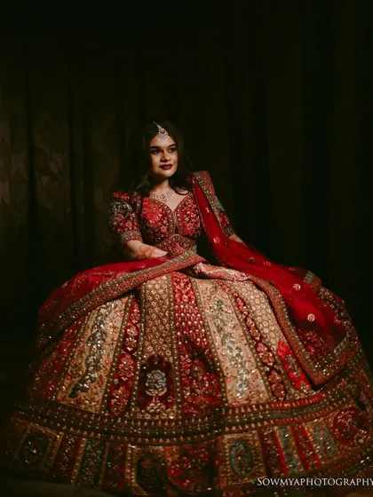 A regal portrait of a bride seated in her elaborate red and gold lehenga. The dark, moody lighting adds to the drama and richness of the image.