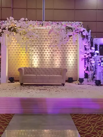 A modern pastel stage setup inside a banquet hall. The backdrop features a textured white panel, accented with an arch of cherry blossom flowers and soft purple lighting.