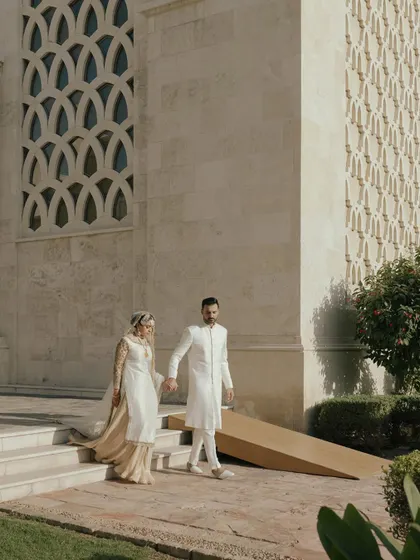 Moin and Roha walking hand-in-hand at the Ismaili Centre in Dubai. Their elegant white outfits are perfect for the serene and modern architectural setting.