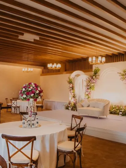 A wider view of the engagement party venue, showing the stage, guest tables with floral centerpieces, and the warm, inviting lighting from the wooden-paneled ceiling.