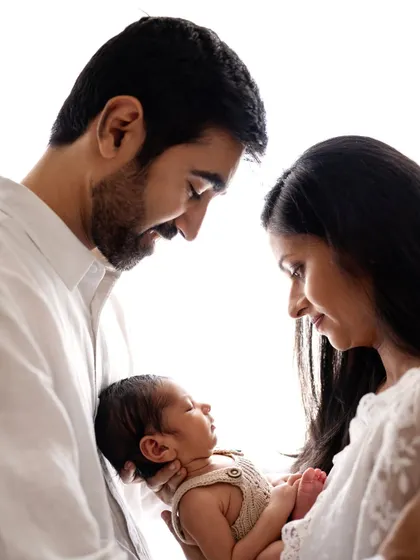 New parents gazing at their sleeping newborn, held between them. The high-key lighting creates a dreamy, angelic feel for this first family portrait.