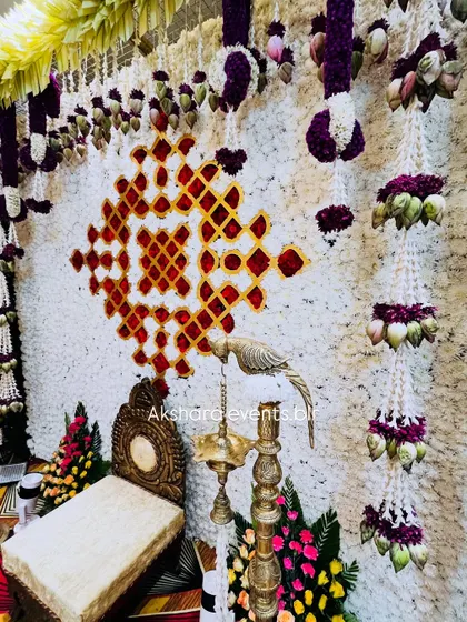 A detailed view of a Haldi or Sreemantham backdrop. The design features a wall of white flowers with a traditional pattern in red, and is adorned with hanging lotus bud garlands.
