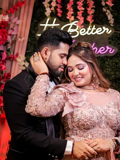 An intimate moment at an engagement party. The groom holds the bride close against a backdrop of flowers and a "Better Together" neon sign, creating a warm, romantic photo.