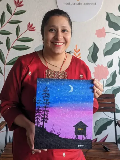 A participant with her beautiful canvas painting of a night sky over a cabin. Our guided workshops make it easy to follow along and create a masterpiece.