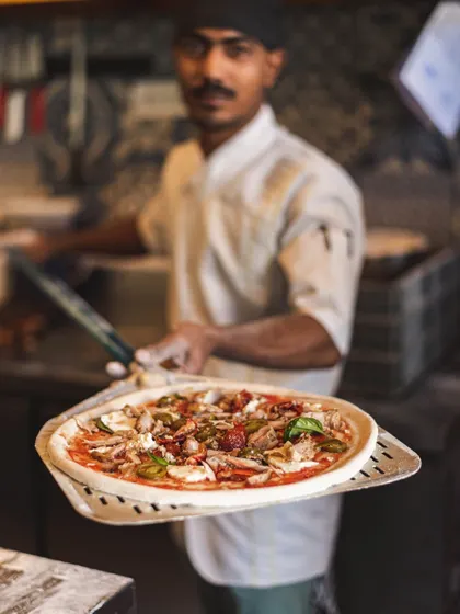 A chef presents a fully topped pizza on a peel, ready for the intense heat of the wood-fired oven. This is the last moment before it transforms into a crispy, bubbly masterpiece.