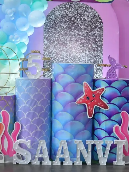 A detailed view of the main stage for a mermaid party. This setup includes custom-printed plinths with scale patterns, coral cutouts, and marquee letters spelling out the name 'SAANVI' on a patch of artificial grass.