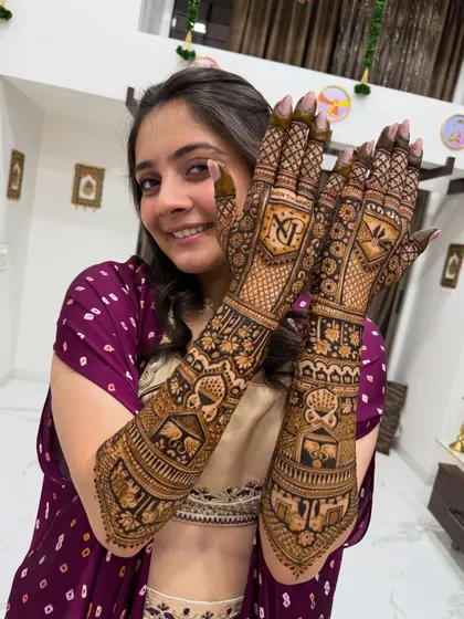 A close-up of a happy bride showcasing her mehndi. This design features the couple's initials "VJ" in a prominent monogram, surrounded by intricate patterns and traditional motifs like a doli and shehnai.