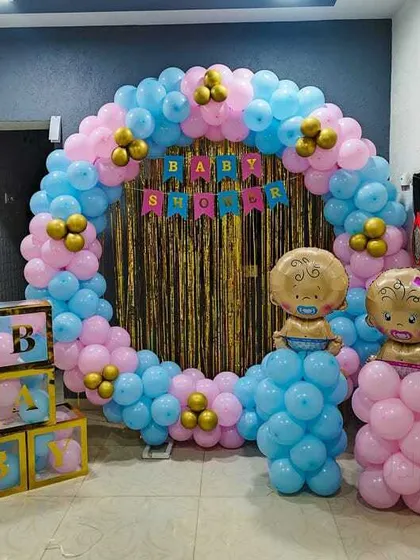 A 'He or She' gender reveal or baby shower setup. A circular balloon arch in pink and blue frames a gold fringe curtain, with 'BABY' blocks and baby-shaped foil balloons.