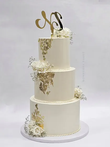 A classic three-tier white and gold leaf wedding cake, adorned with delicate white roses and baby's breath. The custom monogram topper adds a personal touch.