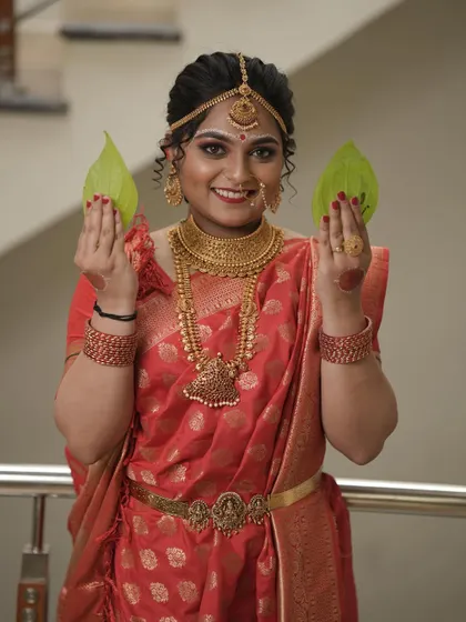 A happy bride holding betel leaves, a beautiful tradition. She is adorned in a complete gold-plated jewellery set, including a matha patti and a statement nath.