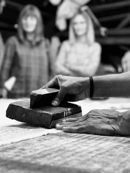 A black and white photograph capturing the essence of our craft. An artisan's hands guide the block, a skill honed over many years, as visitors watch in the background during a studio tour.