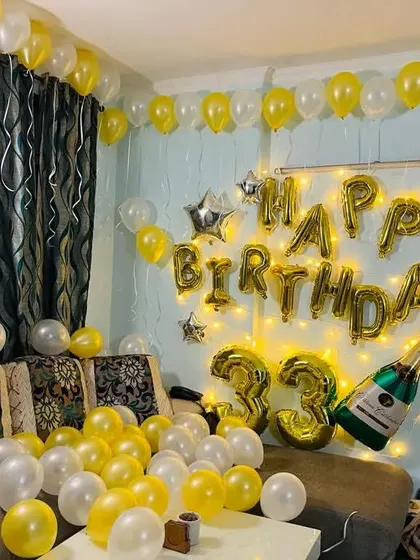 A full view of the yellow and white 33rd birthday room decoration. The couch is surrounded by balloons, and the wall features foil letters and fairy lights.