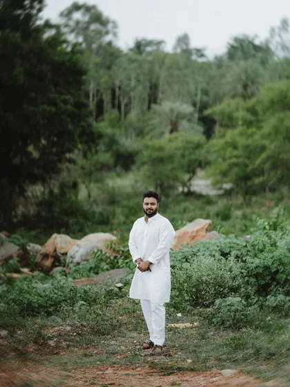 A solo portrait of the groom in a simple white kurta, standing confidently in a lush, green, natural environment.