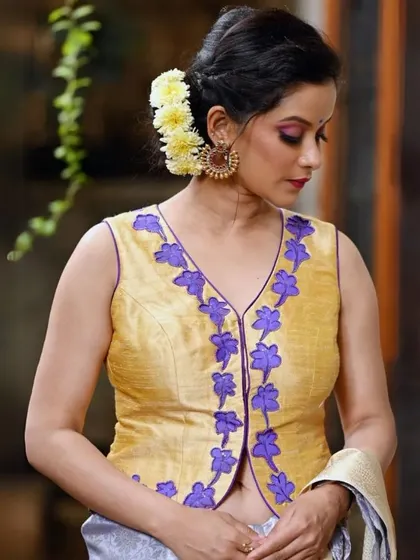 A unique yellow vest-style blouse with purple floral applique is paired with large, traditional gold earrings.