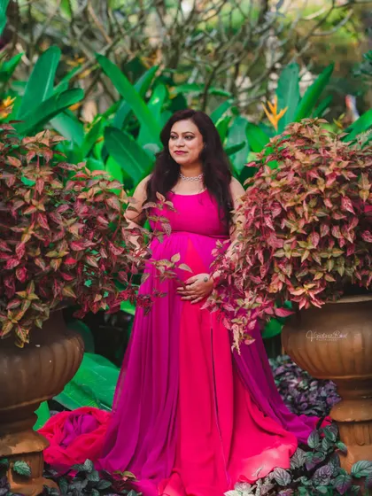 A vibrant portrait of a mother-to-be in a stunning pink and magenta gown, surrounded by colorful foliage in a lush garden setting.