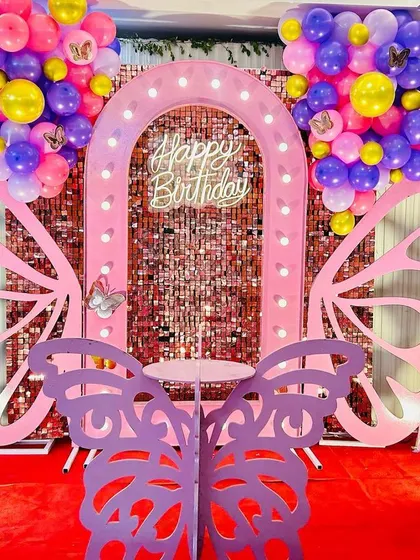 A vibrant butterfly-themed birthday setup with a pink sequin wall. The decor uses bold colors like purple, yellow, and pink in the balloon arrangements and a purple butterfly prop in the foreground.