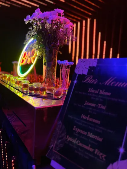 The bar menu is seamlessly integrated into the decor. Here, a custom menu stands next to a neon "BAR" sign and a floral arrangement, with colorful shots lined up and ready for guests.