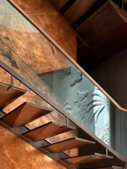 A close-up of the sculptural staircase, showing the cantilevered metal treads and the reflection of the carved wall panel in the glass balustrade.