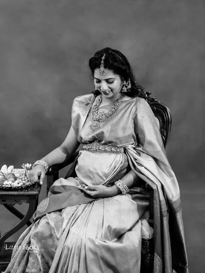 A timeless black and white portrait of an expecting mother in a silk saree. She is seated gracefully, adorned with traditional jewelry, looking down at her baby bump.