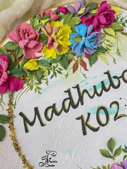 A side view of the "Madhuban" nameplate, showing the contrast between the smooth, hand-lettered names and the highly textured, multi-layered flowers.