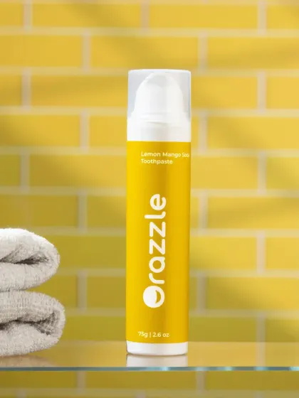 A bathroom lifestyle shot for Orazzle's lemon-mango toothpaste. The yellow tiled background and neatly folded towels create a clean and cheerful scene.