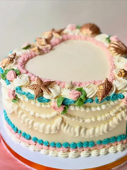 A unique heart-shaped cake with a seaside vintage theme. The classic Lambeth piping is combined with delicate, edible seashells in pink, teal, and gold.