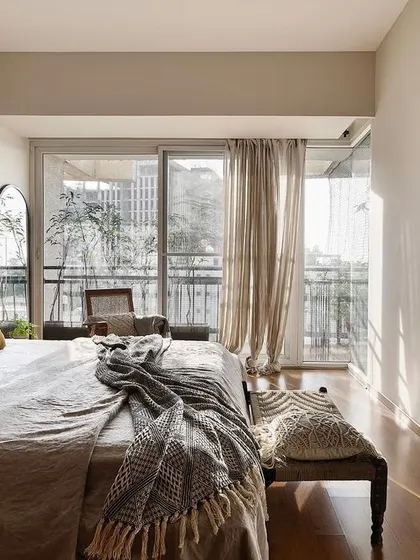 The master bedroom in the 'Reweave Stories' apartment, filled with natural light from the large balcony doors. The design uses a soft, neutral palette and layered textiles to create a comfortable and serene retreat.