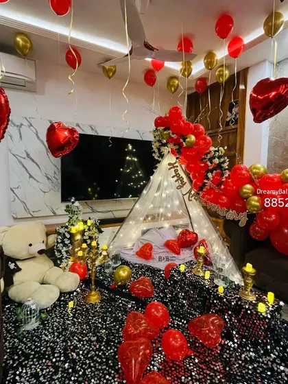 A cozy tent setup in the living room for a special celebration. The floor is covered with a sequin cloth and balloons, with a teddy bear, candles, and a 'Happy New Year' sign creating a festive and intimate space.