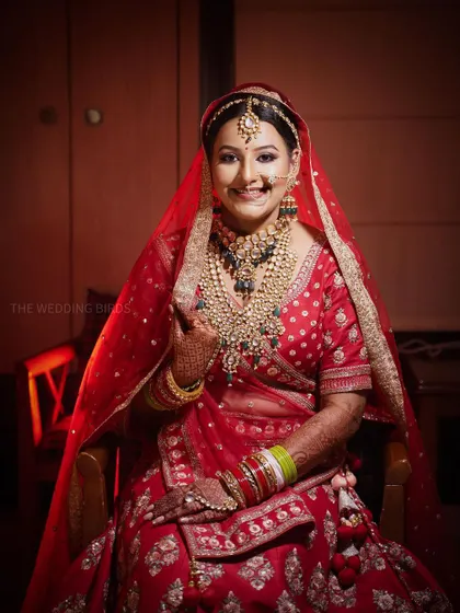 A classic bridal portrait. The bride, adorned in a red lehenga and heavy kundan jewelry, smiles warmly for the camera, looking absolutely timeless.