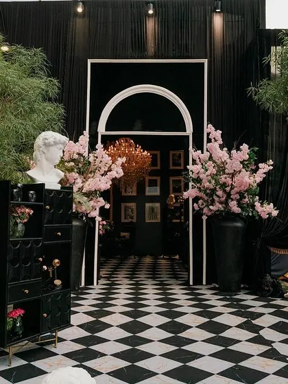 The entrance to a glamorous pre-wedding party. A black-and-white checkered path leads through a modern arched doorway into a room filled with art and elegance, flanked by lush cherry blossom arrangements.