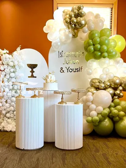 A modern First Communion decoration for a boy, featuring an olive green, white, and gold balloon arch. The setup includes a cake display on textured white plinths against a clean, arched backdrop.