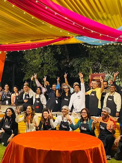 A group photo of all the participants in the corporate cooking challenge, wearing aprons and cheering under a colorful tent. This image captures the spirit of camaraderie and fun that we aim to create.