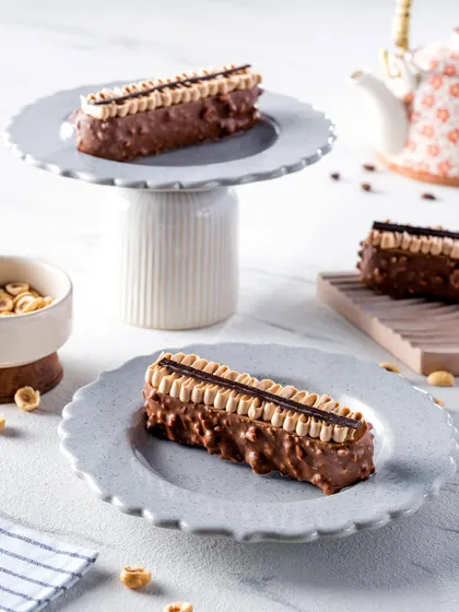 Two hazelnut pastry bars elegantly served on light gray, scalloped plates. The composition, including a small teapot in the background, creates a sophisticated setting for a delightful dessert experience.