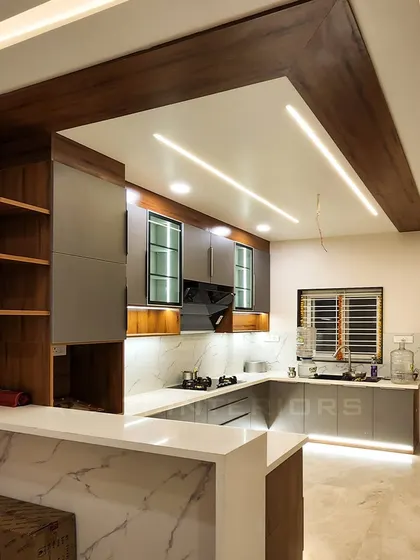 A modern kitchen with a breakfast counter, featuring grey cabinets, a white marble-finish countertop, and warm under-cabinet lighting. The wooden beams on the ceiling add architectural interest.