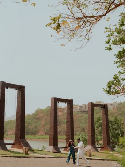 A playful moment captured from a distance, showing the couple dancing near large swings by the lake.