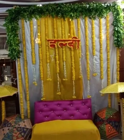 An indoor Haldi seating area with a backdrop of white and yellow drapes, a green foliage border, and hanging marigold strings. The setup is completed with decorative umbrellas.