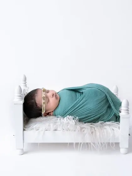 A newborn swaddled in a teal wrap, sleeping soundly in a miniature white bed against a clean, high-key background.