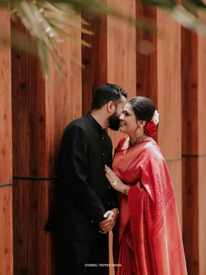 A sweet, stolen moment between Jesna and Antony. The rich red of her saree against the warm wood tones creates a visually stunning and romantic image.