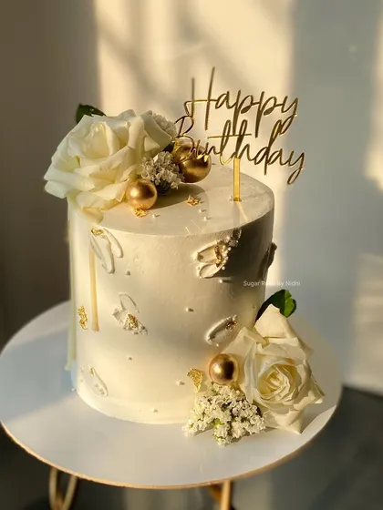A classic white and gold birthday cake. This elegant design is decorated with fresh white roses, gold spheres, and a touch of gold leaf for a luxurious feel.
