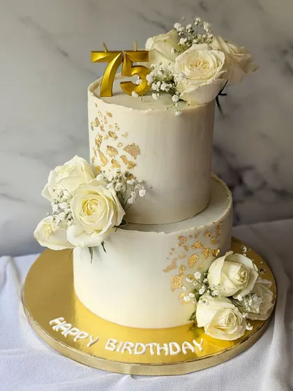 A classic and elegant two-tier cake for a 75th birthday. The design is simple and clean, with a white buttercream finish, gold leaf accents, and fresh white roses.