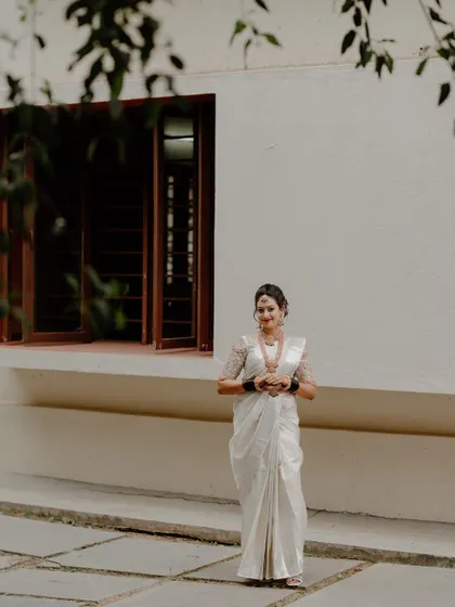 A full-length portrait of a bride in a white saree standing against a simple white wall, creating a clean and minimalist look that emphasizes her traditional attire.