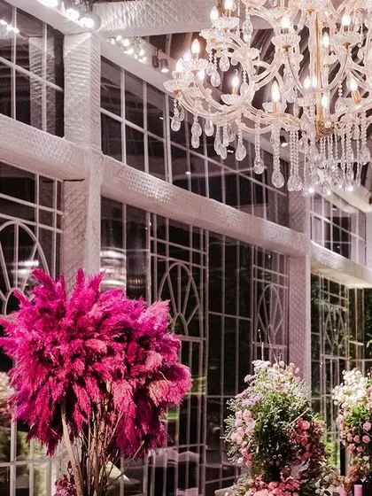 A view of the glasshouse venue, with its soaring ceilings and walls of windows. The space is filled with magnificent crystal chandeliers and dramatic pink floral arrangements.