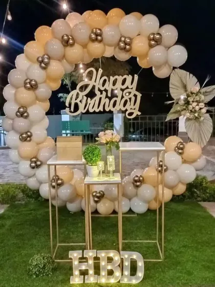 A beautiful outdoor party setup with a balloon ring in neutral tones. The decor includes cake stands and a light-up 'HBD' sign.