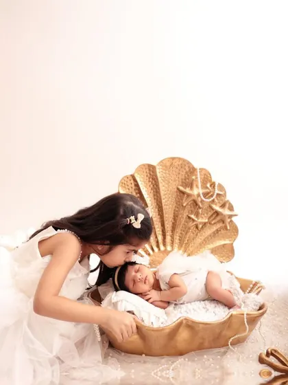 The beginning of a lifelong bond. An older sister gently kisses her newborn sibling, who is resting in a beautiful golden seashell prop. A perfect portrait for celebrating Bhai Dooj.