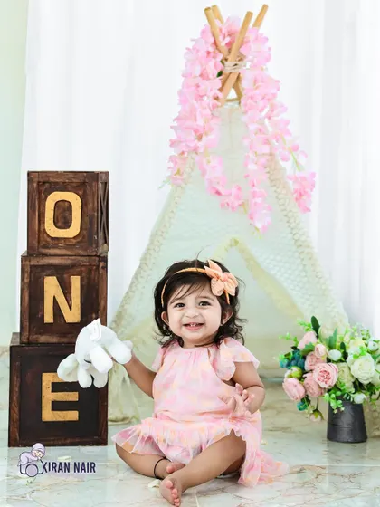 A happy one-year-old girl sits in a studio setup with a teepee and "ONE" blocks. Her bright smile and cute pink dress make this a perfect first birthday portrait.