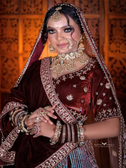 A classic bridal portrait with a confident and direct gaze. The warm lighting enhances the rich maroon color of her velvet outfit.