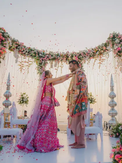 A wide shot of the Varmala ceremony on a beach stage, with the sun setting over the ocean.
