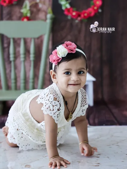 A delightful photo of a baby girl in a white lace dress crawling towards the camera with a sweet smile. The simple studio setup keeps the focus on her happy expression.