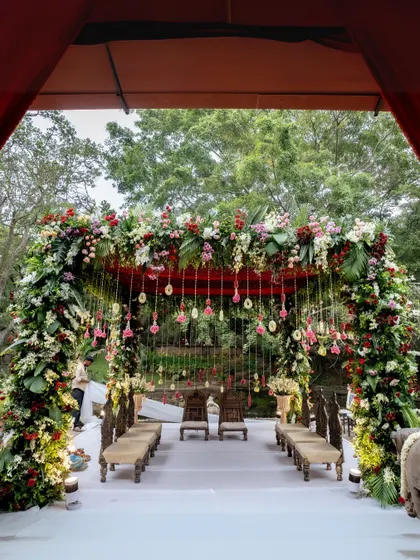 This regal mandap, set beside the serene temple pond, is a masterpiece of floral artistry. A lush archway of red, pink, and white flowers creates a grand entrance to the sacred space, perfect for a traditional wedding ceremony.